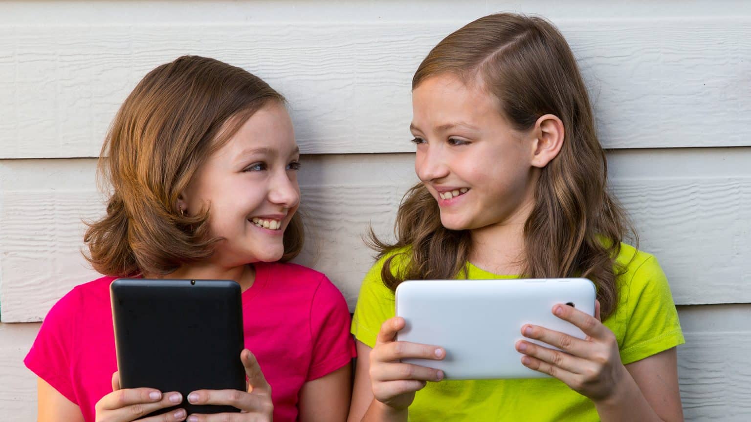 Two happy girls playing with tablet online leaning against a white wall looking each other.