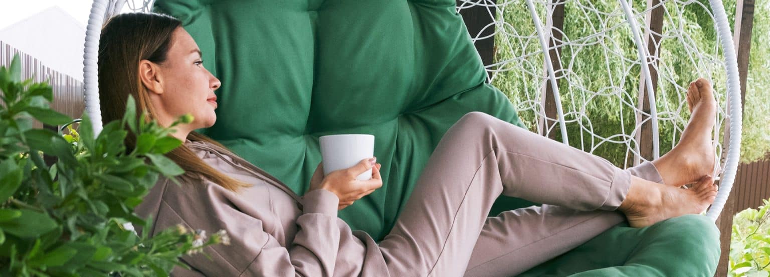 A woman drinking tea or coffee outdoors and sitting in a hammock chair and relaxing in nature. Peaceful and idyllic cozy moments at home or cottage symbolize a restful lifestyle.
