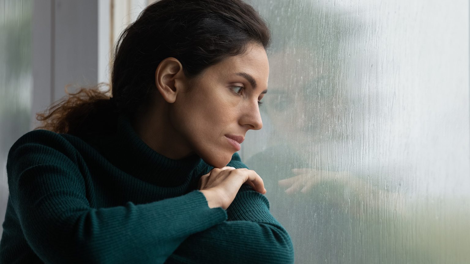 a woman feeling sad looking somberly out the window on a dreary looking day all alone.